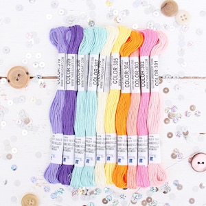 May include: Eight spools of cotton embroidery floss in various colors, including purple, light blue, yellow, orange, pink, and light pink. Each spool has a label with the brand name "Thread Art" and a color number.