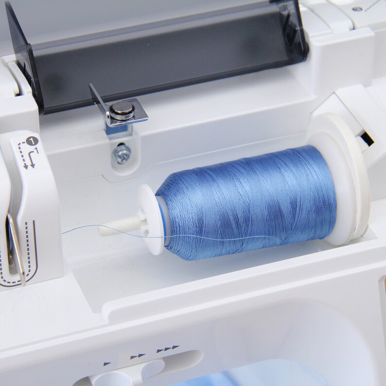May include: A close-up of a sewing machine with a spool of light blue thread. The thread is wound around a white plastic spool holder.