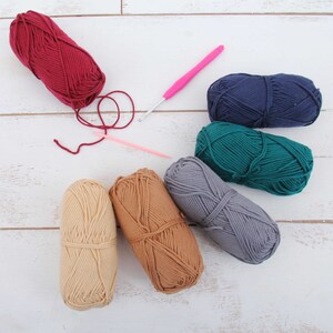 K&ouml;nnte beinhalten: Sechs Kn&auml;uel Garn in verschiedenen Farben, darunter Rot, Blau, Gr&uuml;n, Braun und Beige, mit einem rosa H&auml;kelhaken.