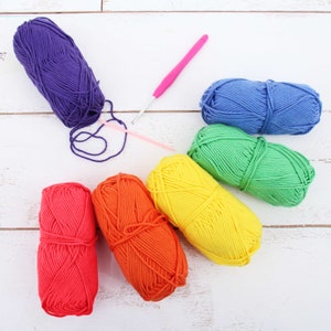 May include: A collection of colorful yarn skeins in red, orange, yellow, green, blue, and purple. Two pink crochet hooks are also present. The yarn is arranged on a white wooden surface, ideal for crafting projects.