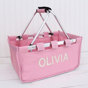 May include: A rectangular pink collapsible basket featuring a silver metal frame and a black padded handle. The front of the basket is embroidered with the name OLIVIA in white capital letters. The basket has a fabric body and metal hinges.
