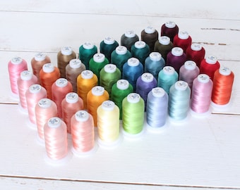 May include: A collection of colorful spools of thread arranged on a white wooden surface. The spools are in various shades, including pink, orange, green, blue, and purple. Each spool has a white base and a label on top.