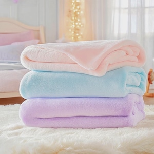 May include: Three soft, plush blankets stacked on top of each other. The top blanket is pink, the middle blanket is light blue, and the bottom blanket is lavender.