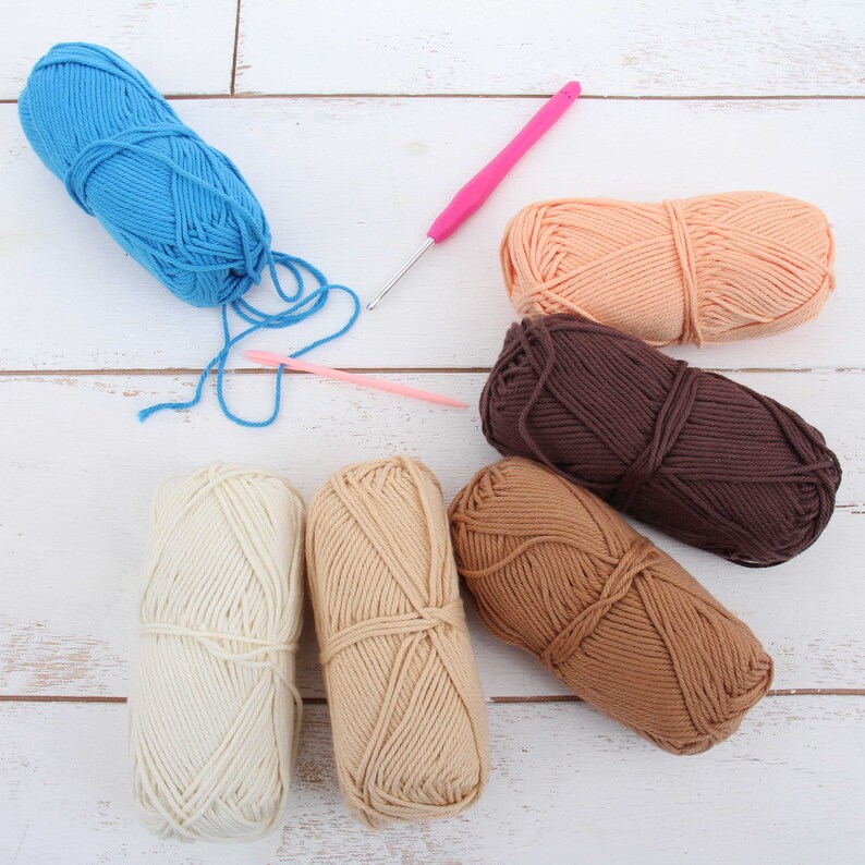 K&ouml;nnte beinhalten: Sechs Garnb&auml;lle in verschiedenen Farben, darunter Blau, Braun, Beige und Pfirsich, liegen auf einer wei&szlig;en Holzoberfl&auml;che. Eine rosa H&auml;kelnadel liegt auf der Oberfl&auml;che.