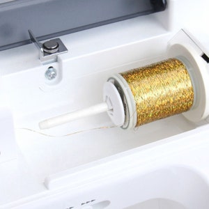 Puede incluir: Primer plano de un carrete de hilo met&aacute;lico dorado en una m&aacute;quina de coser blanca. El hilo est&aacute; enrollado alrededor de un soporte de carrete de pl&aacute;stico blanco y se enhebra a trav&eacute;s de una gu&iacute;a de pl&aacute;stico blanco.