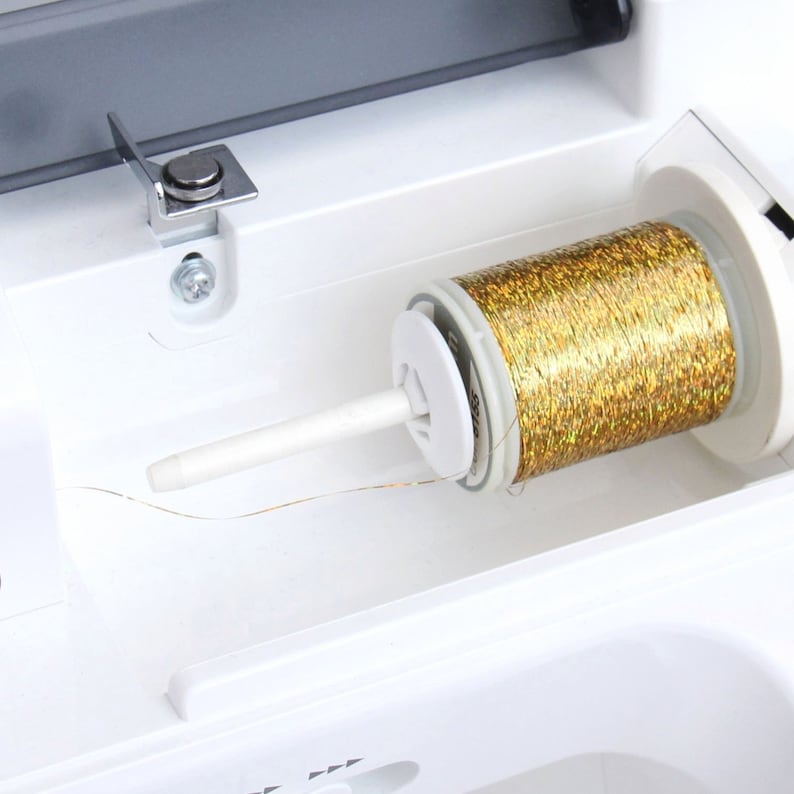 Puede incluir: Primer plano de un carrete de hilo met&aacute;lico dorado en una m&aacute;quina de coser blanca. El hilo est&aacute; enrollado alrededor de un soporte de carrete de pl&aacute;stico blanco y se enhebra a trav&eacute;s de una gu&iacute;a de pl&aacute;stico blanco.