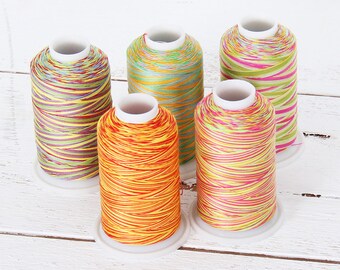 May include: Five spools of colorful embroidery thread, each with a different color combination of stripes. The spools are arranged in a semi-circle on a white wooden surface.