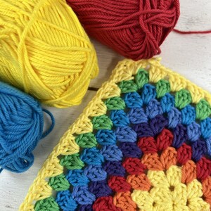 K&ouml;nnte beinhalten: Nahaufnahme eines Regenbogen-H&auml;kel-Grannysquares mit gelbem, rotem, blauem, gr&uuml;nem und violettem Garn. Das Quadrat ist mit einem traditionellen Granny-Square-Muster gearbeitet.