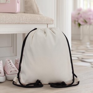 May include: A cream-colored drawstring backpack with black drawstrings. The bag is made of a soft fabric and is sitting on a light-colored floor. Pink sneakers and a pink suitcase are visible in the background.