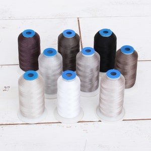 May include: Assortment of spools of thread in various shades of brown, beige, and white. The spools are arranged in a circle on a white wooden surface.