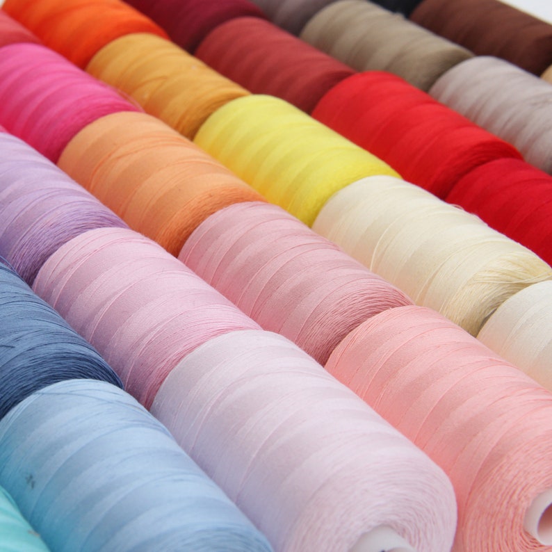 Pode incluir: Uma cole&ccedil;&atilde;o de carret&eacute;is de linha em v&aacute;rias cores, incluindo laranja, amarelo, rosa, azul e verde. Os carret&eacute;is est&atilde;o dispostos em uma fila, com as cores passando gradualmente de uma para a outra.