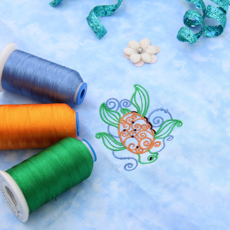 May include: Three spools of embroidery thread in blue, orange, and green. The thread is wound around white spools with blue caps. The spools are on a light blue fabric with a decorative embroidered turtle design.