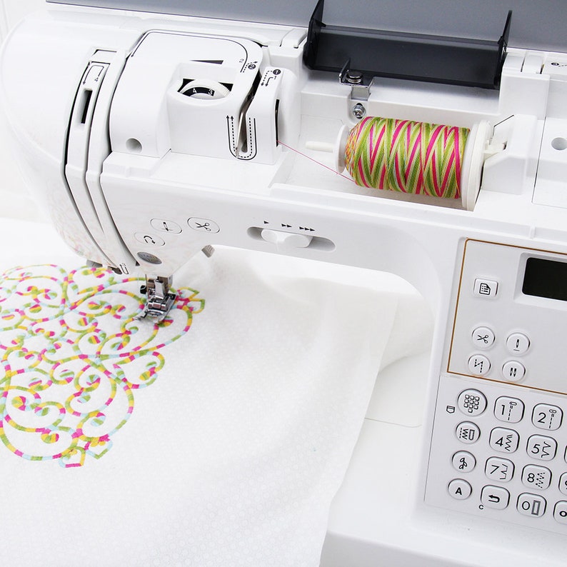 May include: A white sewing machine with a spool of green, pink, and yellow thread in the bobbin winder. The machine is embroidering a white fabric with a colorful floral design.