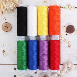 May include: Eight spools of thread in various colors, including black, white, yellow, orange, green, blue, purple, and red. The spools are arranged in two rows of four, with the spools in each row alternating in color.