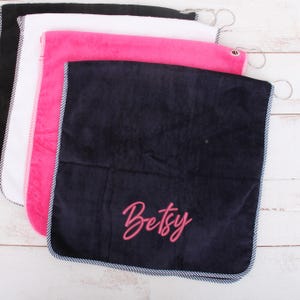 Könnte beinhalten: Ein Stapel von vier quadratischen Golfhandtüchern in Schwarz, Weiß, Pink und Marineblau. Das marineblaue Handtuch ist mit dem Namen "Betsy" in Pink bestickt. Jedes Handtuch hat einen gestreiften Rand und einen Metallring zum Aufhängen.