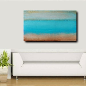 May include: Abstract painting with horizontal stripes of light brown, blue, and white. The painting is hanging on a white wall above a white couch.