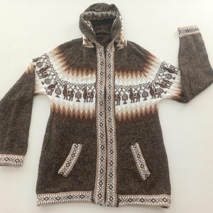 Könnte beinhalten: Ein brauner Kapuzenpullover mit Reißverschluss und zwei Vordertaschen. Der Pullover hat ein weißes, braunes und beiges geometrisches Muster mit Lama- und Vogelmotiven. Die Manschetten und der Saum haben einen gemusterten Rand.