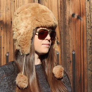 May include: A woman wearing a brown faux fur trapper hat with ear flaps and pom-poms. She is also wearing aviator sunglasses with a green frame and brown lenses. The background is a wooden wall.