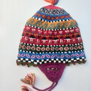 Könnte beinhalten: Eine bunte Strickmütze mit Ohrenklappen, die komplexe geometrische und figürliche Muster in Rot, Blau, Grün, Orange und Weiß aufweist. Die Mütze ist mit einem mehrfarbigen Wollbommel versehen und hat geflochtene Bänder mit Quasten an den Enden.