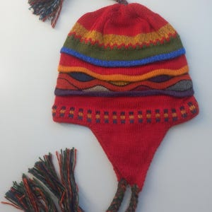 Könnte beinhalten: Eine rote Strickmütze mit Ohrenklappen, einer geflochtenen Quaste oben und zwei geflochtenen seitlichen Zöpfen. Die Mütze zeigt horizontale Streifen in Gelb, Olivgrün, Blau und Orange sowie wellenförmige Muster in Violett und Dunkelrot.