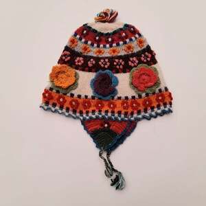 May include: A colorful, hand-knitted hat with a floral design. The hat features a cream-colored base with vibrant patterns in red, orange, blue, and green. It has ear flaps and a tassel, and is adorned with several crocheted flowers.