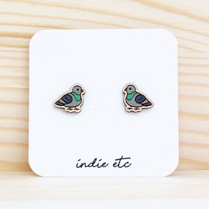 May include: A pair of wooden earrings featuring a small, gray and green pigeon design. The earrings are on a white card with the text "indie etc".