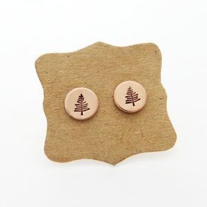 May include: A pair of small, round, rose gold earrings with a tree design. The earrings are displayed on a brown card.