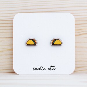 May include: A pair of wooden earrings shaped like tacos. The earrings are painted yellow, green, and brown. The earrings are on a white card with the text "indie etc" printed on it.