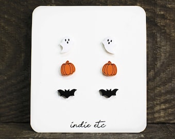 May include: A set of six Halloween-themed stud earrings. The earrings feature a white ghost, an orange pumpkin, and a black bat. The earrings are displayed on a white card with the text "indie etc".