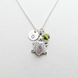 May include: Silver necklace with a turtle charm, a green crystal bead, and a silver disc charm with the letter "O" engraved on it.
