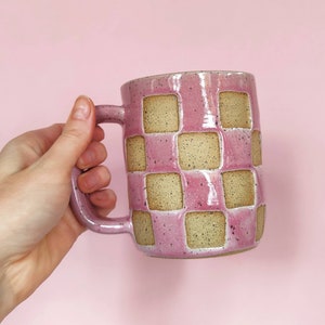 Pode incluir: Uma caneca de cerâmica rosa com um padrão xadrez em cor marrom claro. A caneca tem uma alça e é salpicada de manchas marrons.