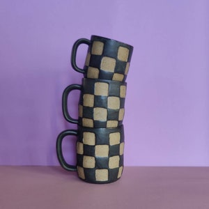 Puede incluir: Tres tazas de cer&aacute;mica negra apiladas una encima de la otra. Cada taza tiene un patr&oacute;n de cuadros con cuadrados de color beige.