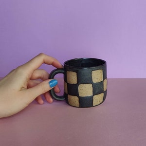 Puede incluir: Una taza de cer&aacute;mica negra con un patr&oacute;n de cuadros en beige. La taza tiene un asa y es parcialmente visible en la mano de una persona.