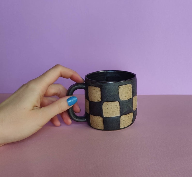 Puede incluir: Una taza de cer&aacute;mica negra con un patr&oacute;n de cuadros en beige. La taza tiene un asa y es parcialmente visible en la mano de una persona.
