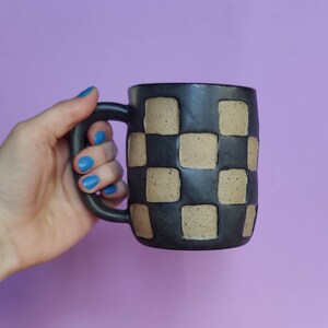 Puede incluir: Una taza de cer&aacute;mica negra con un patr&oacute;n de cuadros en marr&oacute;n. La taza tiene un asa redondeada.