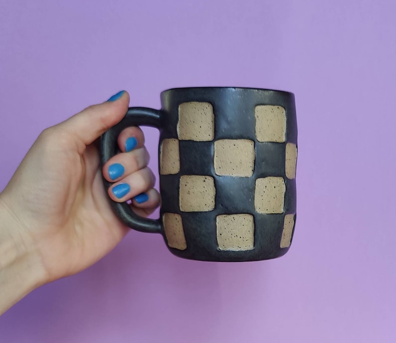 Puede incluir: Una taza de cer&aacute;mica negra con un patr&oacute;n de cuadros en marr&oacute;n. La taza tiene un asa redondeada.