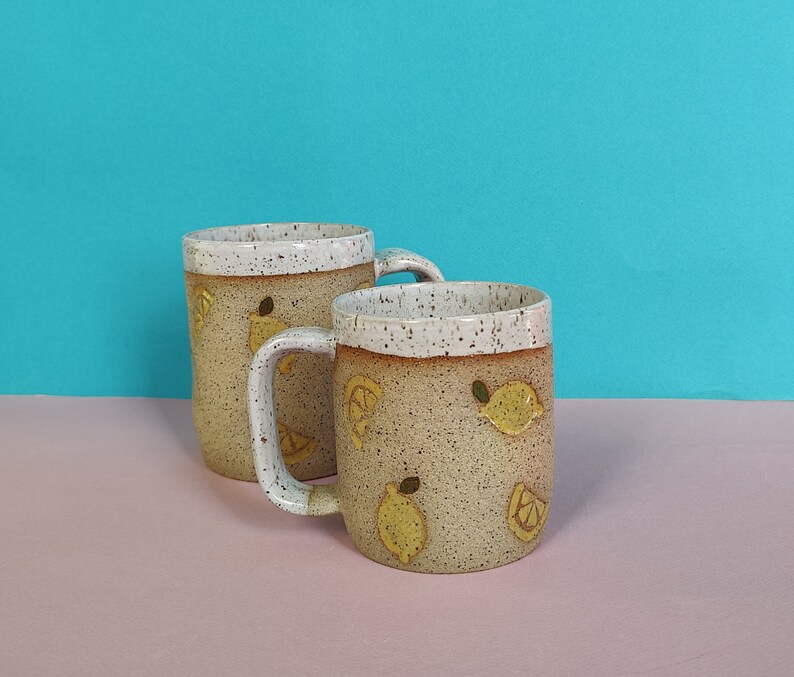 May include: Two ceramic mugs with a speckled brown and white glaze. The mugs have a lemon design with yellow accents.