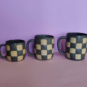 Puede incluir: Tres tazas de cer&aacute;mica con un patr&oacute;n de cuadros en negro y marr&oacute;n. Las tazas son de diferentes tama&ntilde;os.
