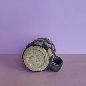 Puede incluir: Una taza de cer&aacute;mica negra y marr&oacute;n con un patr&oacute;n a cuadros. La taza tiene un asa y un esmalte moteado.