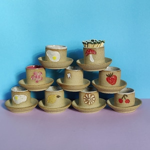 May include: A set of ten ceramic cups with various designs, including a flower, a tooth, a strawberry, a mushroom, and a cherry. The cups are arranged in a pyramid shape on a light purple surface.