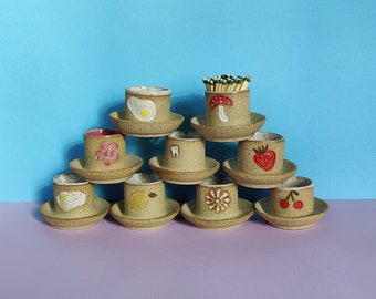 May include: A set of ten ceramic cups with various designs, including a flower, a tooth, a strawberry, a mushroom, and a cherry. The cups are arranged in a pyramid shape on a light purple surface.