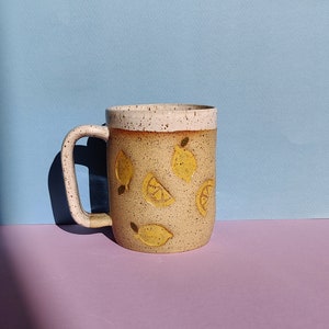 May include: A ceramic mug with a white rim and a speckled brown glaze. The mug features a yellow lemon design with whole lemons and lemon slices.