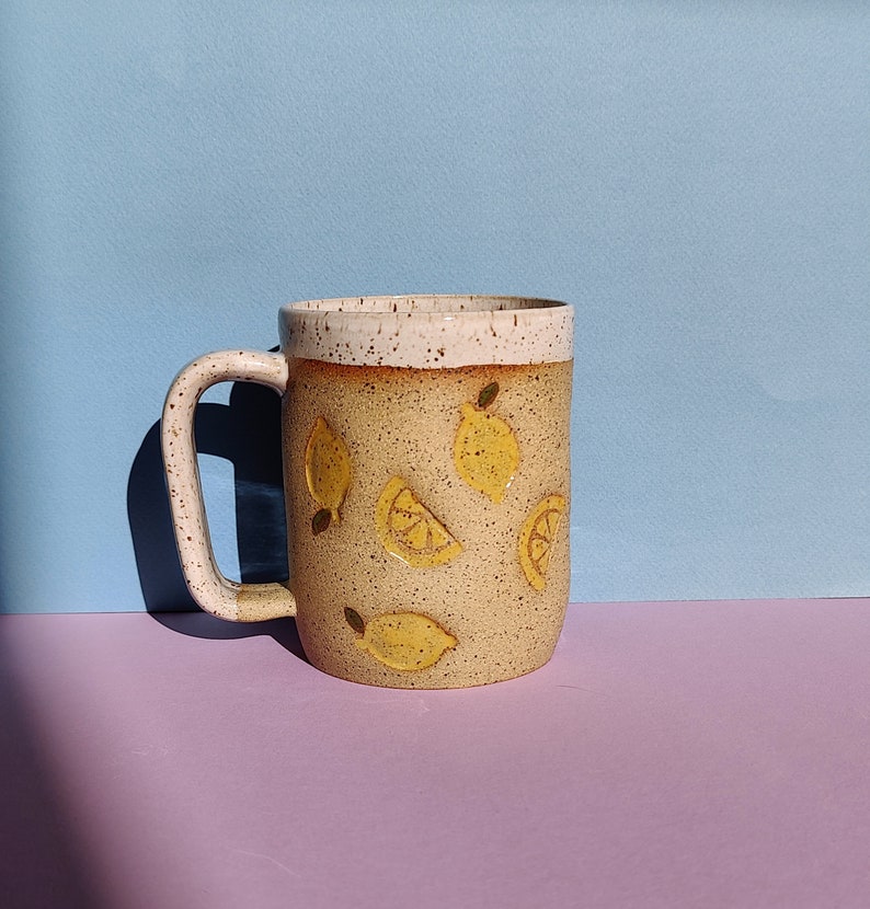 May include: A ceramic mug with a white rim and a speckled brown glaze. The mug features a yellow lemon design with whole lemons and lemon slices.