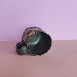 Puede incluir: Una taza de cer&aacute;mica verde oscuro con un patr&oacute;n geom&eacute;trico de cuadrados beige. La taza est&aacute; sobre una superficie rosa claro.