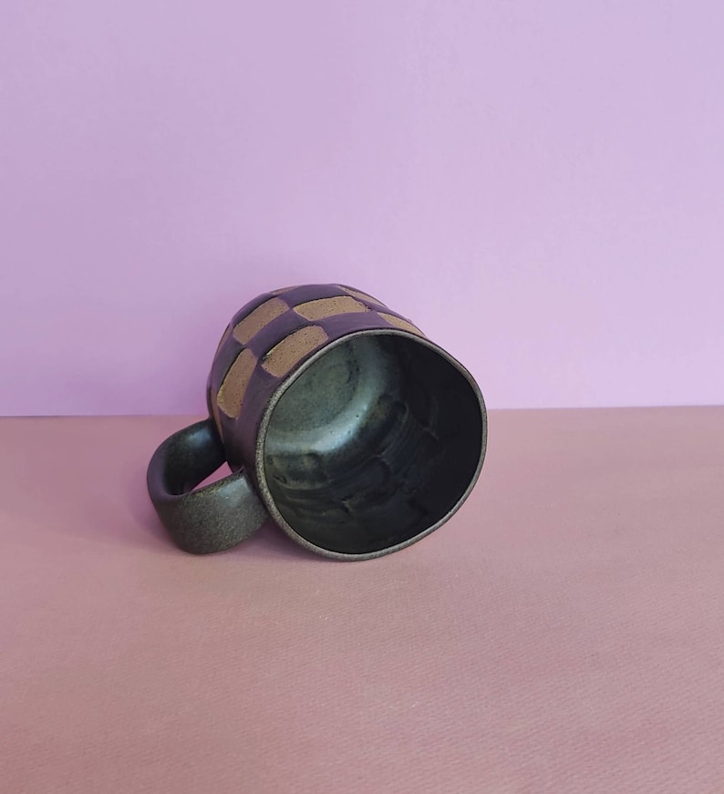Puede incluir: Una taza de cer&aacute;mica verde oscuro con un patr&oacute;n geom&eacute;trico de cuadrados beige. La taza est&aacute; sobre una superficie rosa claro.