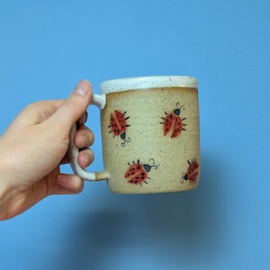 May include: A beige ceramic mug with a white rim and a handle. The mug is decorated with a pattern of red ladybugs.