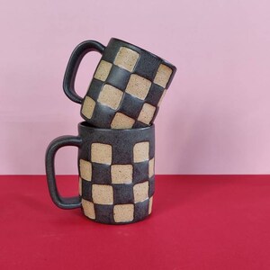 Puede incluir: Dos tazas de cer&aacute;mica negra con un patr&oacute;n de cuadros en beige y negro. Las tazas est&aacute;n apiladas una encima de la otra.