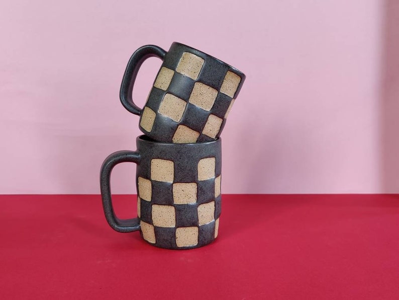 Puede incluir: Dos tazas de cer&aacute;mica negra con un patr&oacute;n de cuadros en beige y negro. Las tazas est&aacute;n apiladas una encima de la otra.