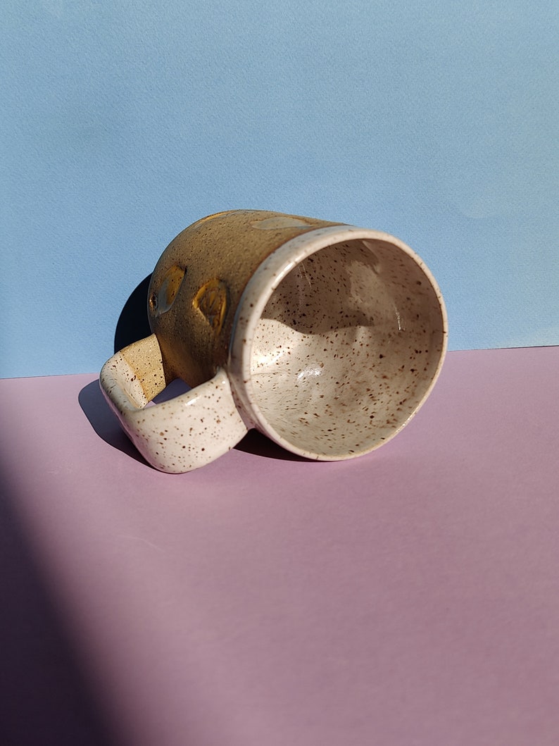 May include: A speckled brown and white ceramic mug with a yellow and brown design. The mug is tilted on its side, showing the inside of the mug.