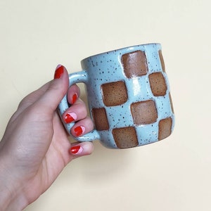 May include: A light blue ceramic mug with a brown checkered pattern. The mug has a speckled glaze and a rounded handle.